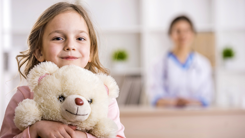 Ein Foto. Im Vordergrund hält ein junges Kind mit langen, hellbraunen Haaren und einem leichten Lächeln einen cremefarbenen Teddybären fest an seine Brust. Das Kind trägt ein hellrosa langärmeliges Oberteil, und der Teddybär hat ein einfach gesticktes Lächeln und eine braune Nase. Der Hintergrund ist unscharf und zeigt eine Person in einem weißen Kittel mit einem Stethoskop, was auf eine klinische Umgebung oder ein Büro mit weißen Regalen und kleinen grünen Pflanzen hindeutet. Die Beleuchtung ist hell und gleichmäßig, wodurch das Kind und der Bär als zentraler Fokus hervorgehoben werden.