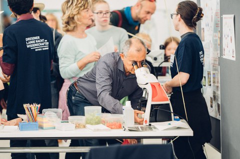 A photo of a person looking through a microscope. In the background, several people are talking.