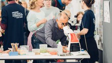 A photo of a person looking through a microscope. In the background, several people are talking.