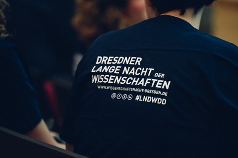 A photo of the back of a person. They are wearing a T-shirt with a print: „DRESDNER LANGE NACHT der WISSENSCHAFTEN“ under that, the internet address: „www.wissenschaftsnacht-dresden.de“. Below that, four logos of Instagram, facebook, X (former Twitter) und LinkenIn, besides that there’s the Hashtag: #LNDWDD.