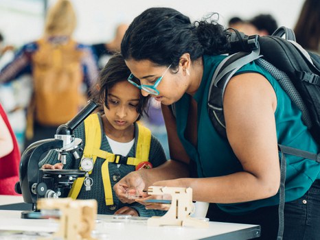 A photo of a person with their child at an old microscope, holding a glass container.