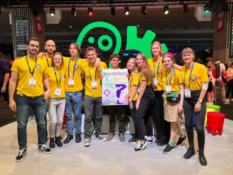 A group photo. 11 young people pose together. All are wearing the same yellow t-shirts. The person in the middle holds a poster that says "WunderBand"