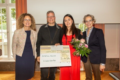 ein Gruppenfoto. Für die Menschen posieren drei Damen und ein Herr. Die jüngste Dame hält Blumen und einen großen Scheck