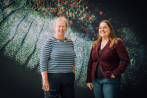 two people standing next to each other smiling. Behind them, there is a colorful background of a painting.