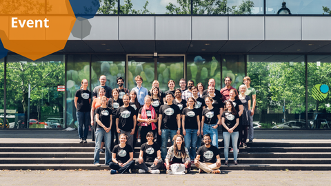 A group photo. About 30 people are organized in four rows on the stairs in front of a building. Most of the people wear matching t-shirts. The building has glass walls that reflect the green trees and blue sky.