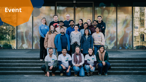 A photo with graphical elements. A group photo of 20 people standing in four rows on stairs in front of a building. The people are looking into the camera and smiling. People in the front row have visible nametags clipped to their identical grey t-shirts. In the background, a colorful glass door is visible. In the upper left corner, there are colorful octagons with a word "Event" written in them.