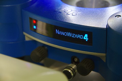 Ein Foto. Der Hintergrund ist ein metallblauer Kreis. In der Mitte befindet sich ein schwarzes Display mit leuchtend blauem Text: „NANOWIZARD 4“. Links daneben sind ein rotes und ein blaues Licht.