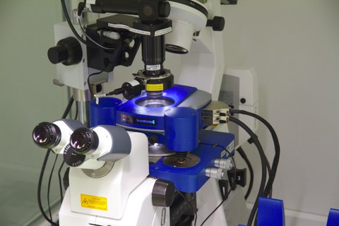 A photo. The background is light grey. A white and blue microscope with multiple lenses is in the center. There is text: "NANOWIZARD 4". There are two black knobs, eight cables, and three white tubes.
