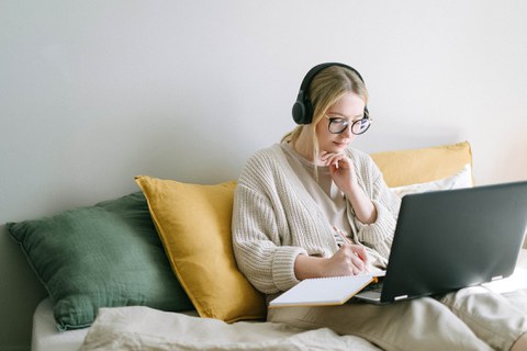 Blonde, junge Frau sitzt auf einem Bett mit einem Laptop auf dem Schoß.