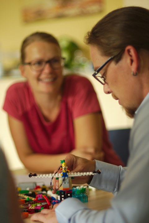 Im Vordergrund baut eine männliche Person mit Brille und Bart Lego zusammen, das vor ihm auf dem Tisch liegt. Verschwommen im Hintergrund sitzt ebenfalls am Tisch eine lächelnde Frau mit blondem Haar und Brille.