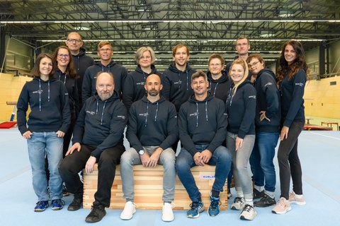 Foto Gruppe von Personen in einer Sporthalle, mehrere sitzend, dahinter mehrere stehend, alle in dunkelblauen Kapuzenpullovern