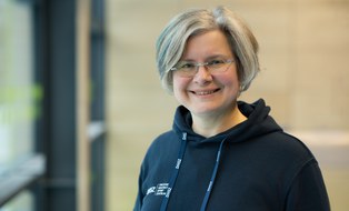Foto: Frau mit kinnlangen grau-blonden Haaren und Brille, im dunkelblauen Kapuzenpullover mit DHSZ-Logo