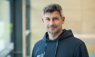 Foto: Mann mit kurzen grau-schwarzen Haaren, im dunkelblauen Kapuzenpullover mit DHSZ-Logo