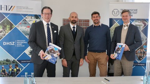 Foto 4 Männer stehen hinter einem Tisch zwischen Bannern mit DHSZ-Bildern und Logos
