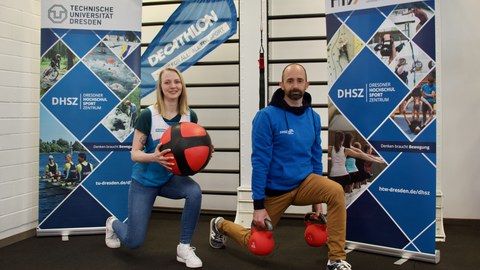 Foto: eine Frau mit einem Medizinball und ein Mann mit roten Kettlebells posieren zwischen Banners des Dresdner Hochschulsportzentrums