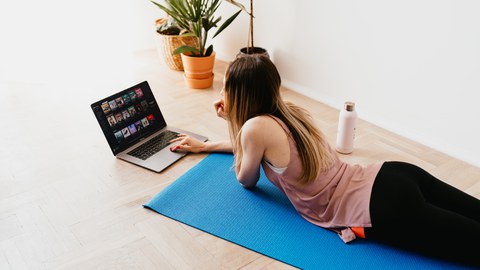 Foto: Junge Frau in Sportkleidung liegt auf einer Yogamatte vor einem Laptop