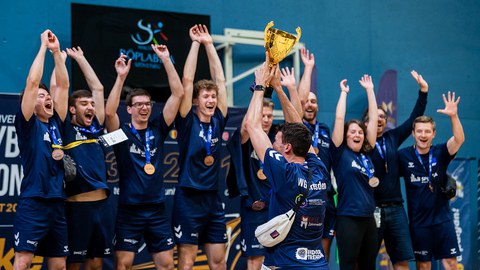 Foto: Sportler in dunkelblauen T-Shirts jubeln über einen bronzenen Pokal
