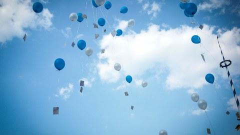 Das Foto zeigt zahlreiche graue und blaue Luftballons. Sie steigen in den blauen Himmel auf. An ihnen sind Postkarten befestigt. Am rechten Bildrand erkennt man zudem einen Maibaum.