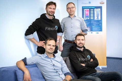 zwei Männer sitzen auf einem Sofa, zwei weitere stehen hinter ihnen, zwei der Personen tragen einen Pullover mit Aufdruck "Flexora", im Hintergrund eine Rollup mit dem Schriftzug "Flexora"