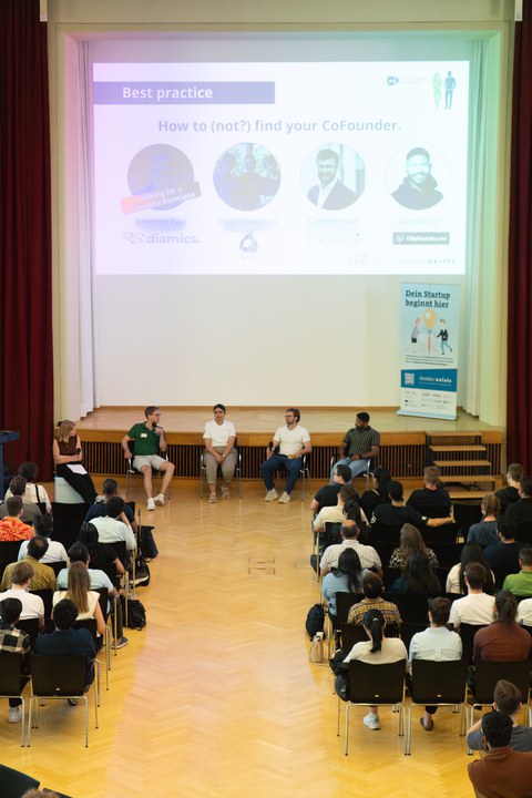 Four people are seated in front of the stage, participating in a panel discussion; the audience is seated in front of them 