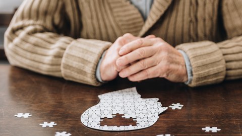 Foto eines älteren Mannes. Vor ihm liegt ein Puzzle, das einen menschlichen Kopf darstellt. Im Bereich des Gehirns fehlen Teile, die darum verstreut liegen.