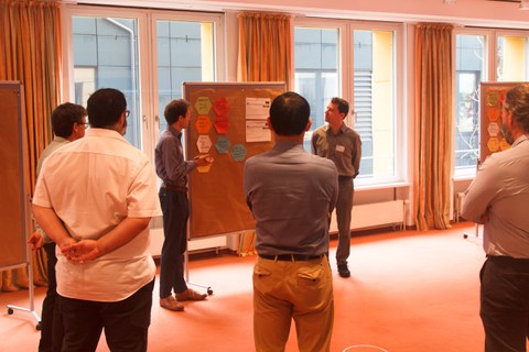 Participants of the FUTURE STUDIO are standing around a pinboard, observing the results that have been developed there.
