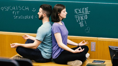 Zwei Studierende sitzen Rücken an Rücken im Schneidersitz in einer meditierenden Armhaltung auf dem Pult vor der Tafel im Hörsaal und haben die Augen geschlossen.