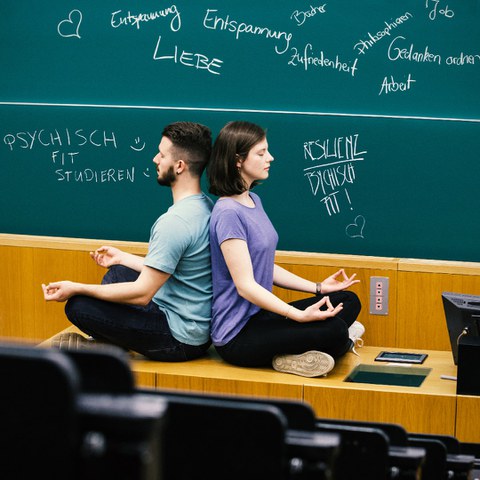 Zwei Studierende sitzen Rücken an Rücken im Schneidersitz in einer meditierenden Armhaltung auf dem Pult vor der Tafel im Hörsaal und haben die Augen geschlossen.