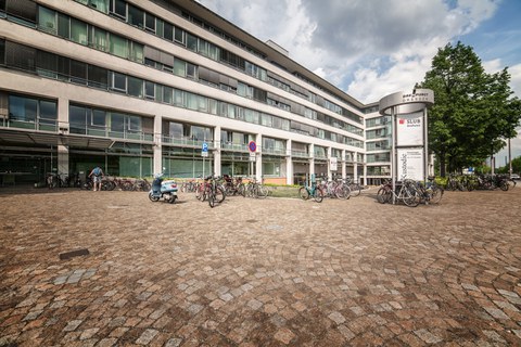 Der Eingang vom Bürogebäude Zellescher Weg. Zu sehen sind die große Fensterfassade des Gebäudes, viele Fahrräder, ein Motorroller, ein Baum und eine Informationssäule.