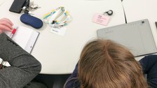 Blick von oben auf einen Konferenztisch mit Tablet und Schreibsachen, daneben ein Besucherschild "Universitätsschule Dresden" und "Forschungsstelle Universitätsschule ForUS"