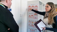 Eine Projektmitarbeiterin erklärt einem Schulleiter die sogenannten Lernhäuser auf einem Plakat in der Materialbibliothek der Universitätsschule Dresden.