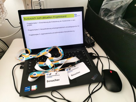 Ein Laptop zeigt die Folie "Austausch zum aktuellen Projektstand". Auf der Tastatur liegen Besucher-Schilder der Universitätsschule Dresden.