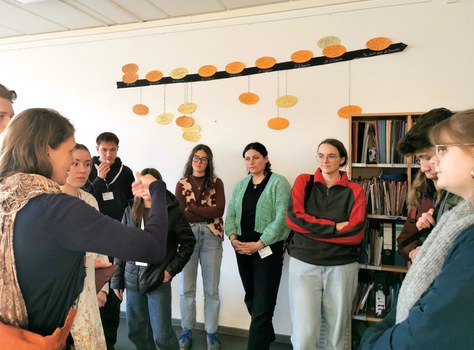 10 Studierende bei der Schulführung in einem Lernraum. Sie stehen vor einem Materialregal und unter einem Zeitstrahl. Sie schauen zur Projektmitarbeitern, die erklärt.