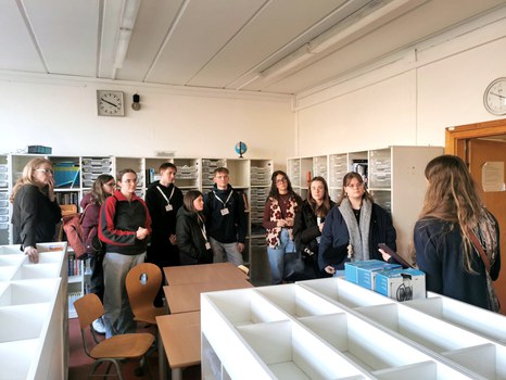 8 Studierende und zwei Projektmitarbeiterinnen in der Materialbibliothek für die Jahrgangsstufen 7-10 der USD. An den Wänden stehen Regale mit transparenten offenen Schubkästen, davor weitere Regale mit Kästen darauf, dazwischen Tische und Stühle.