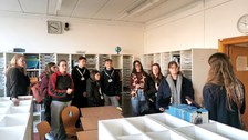 8 Studierende und zwei Projektmitarbeiterinnen in der Materialbibliothek für die Jahrgangsstufen 7-10 der USD. An den Wänden stehen Regale mit transparenten offenen Schubkästen, davor weitere Regale mit Kästen darauf, dazwischen Tische und Stühle.