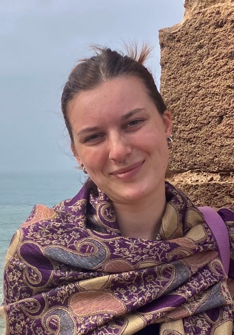 Eine junge Frau schaut in die Kamera. Im Hintergrund ist das Meer und eine alte Steinmauer zu sehen.