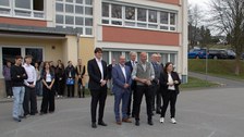 Vor einer Plattenbauschule steht eine Gruppe Erwchachsener, dahinter vereinzelte Schülerinnen und Schüler.