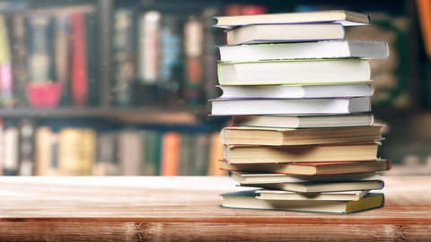 Foto: Ein Bücherstapel liegt auf einem Tisch. Im Hintrgrund ist ein Bücherregal.