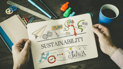 Ein Arbeitsplatz, über dem zwei Hände ein Notizbuch halten mit einer Skizze zum Thema "Sustainabilty".