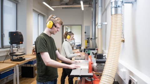 Zwei Studierenden stehen an Schleifmaschinen und schleifen Holz