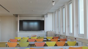 Raum mit gelben und orangenen Stuhlreihen und einer digitalen Tafel