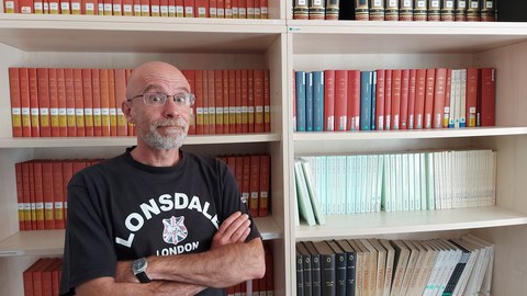 Florent Cygler steht vor einem Bücherregal in der Bibliothek der FOVOG. Er trägt ein schwarzes T-Shirt und verschränkt die Arme.
