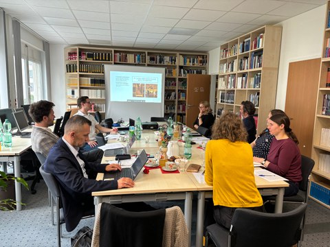 Blick in die Bibliothek der FOVOG. Viele Menschen sitzen an einem Tisch, im Hintergrund ist eine Präsentation zu sehen. 