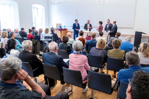 Blick von hinten über die Stuhlreihen eines gut gefüllten Tagungsraums auf ein Stehpodium, an dem fünf Personen in Geschäftskleidung stehen und sprechen.