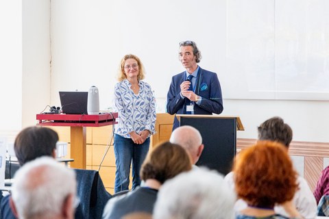 Mirko Breitenstein spricht mit einem Mikrofon an einem Rednerpult vor Publikum, während Gaby Lindenmann-Merz lächelnd neben ihm steht.