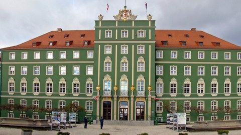 Zu sehen ist die Hauptfront des Rathauses von Stettin mit dem Wappen der Stadt auf dem Giebel. Auf dem Platz sind zwei Zierbrunnen und kleine Bäume.