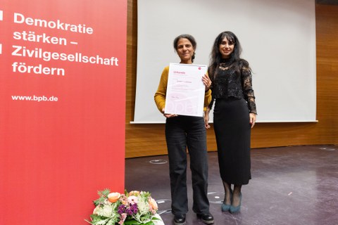 Links ein Banner, rechts zwei Personen. Die Person neben dem Banner hält eine Urkunde. 