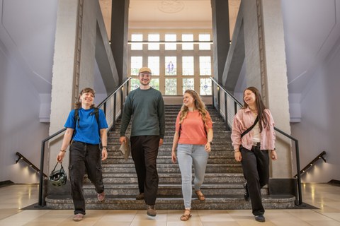 Photo: Four students are walking down a wide staircase, laughing; a large window can be seen in the background.