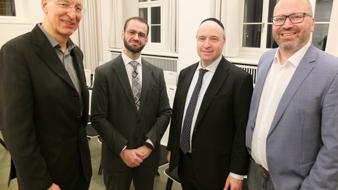 Das Bild zeigt 3 Männer neben einander stehend. Sie haben einen freundlichen bis neutralen Gesichtsausdruck. Alle tragen Anzüge, der zweite von rechts trägt zu dem eine Kippa.