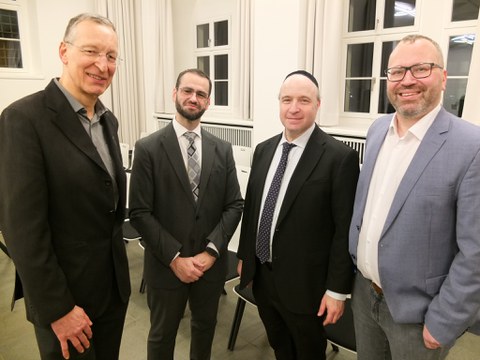 Das Bild zeigt 3 Männer neben einander stehend. Sie haben einen freundlichen bis neutralen Gesichtsausdruck. Alle tragen Anzüge, der zweite von rechts trägt zu dem eine Kippa.